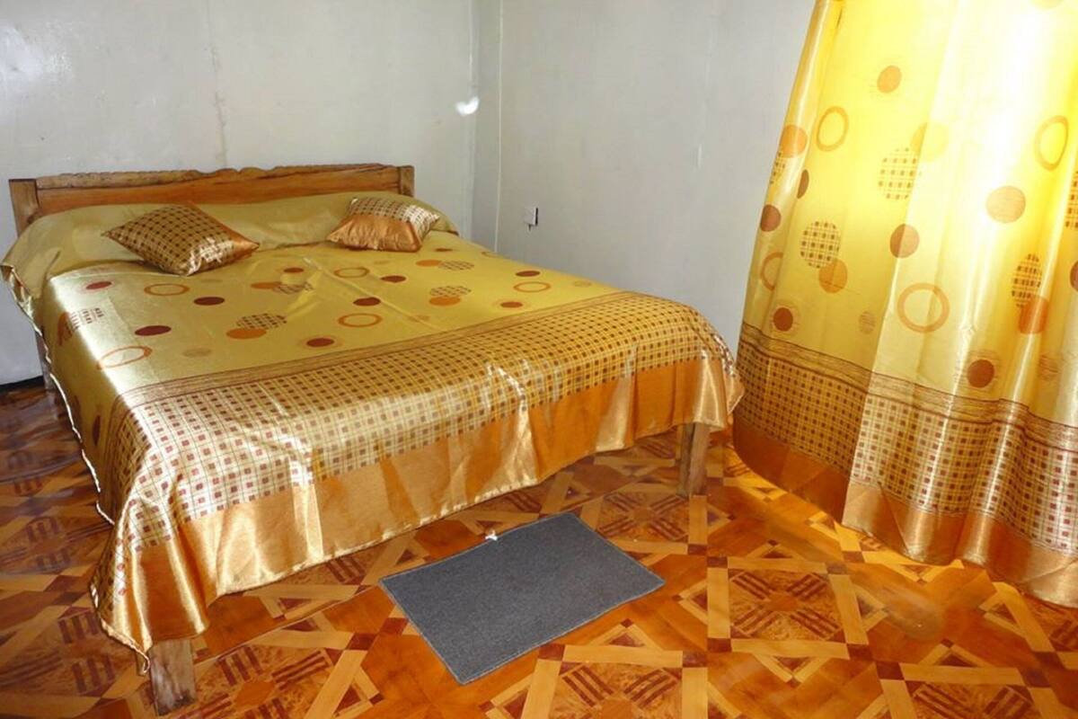 A single bed with a patterned bedspread and pillows, a wooden headboard, and a patterned floor.