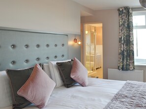 A hotel room with a tufted headboard, a large bed with multiple pillows, a bathroom visible through an open door, and a window with floral curtains.