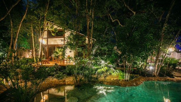 A nighttime view of a house surrounded by lush greenery and a clear pool. - Nahouse Jungle Lodges (Tulum)