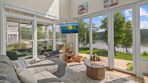 A spacious living room with a large window overlooking a lake, a comfortable gray sofa, and a flat-screen TV mounted on the wall. - Bow-wow on The River #5 (Ottawa)