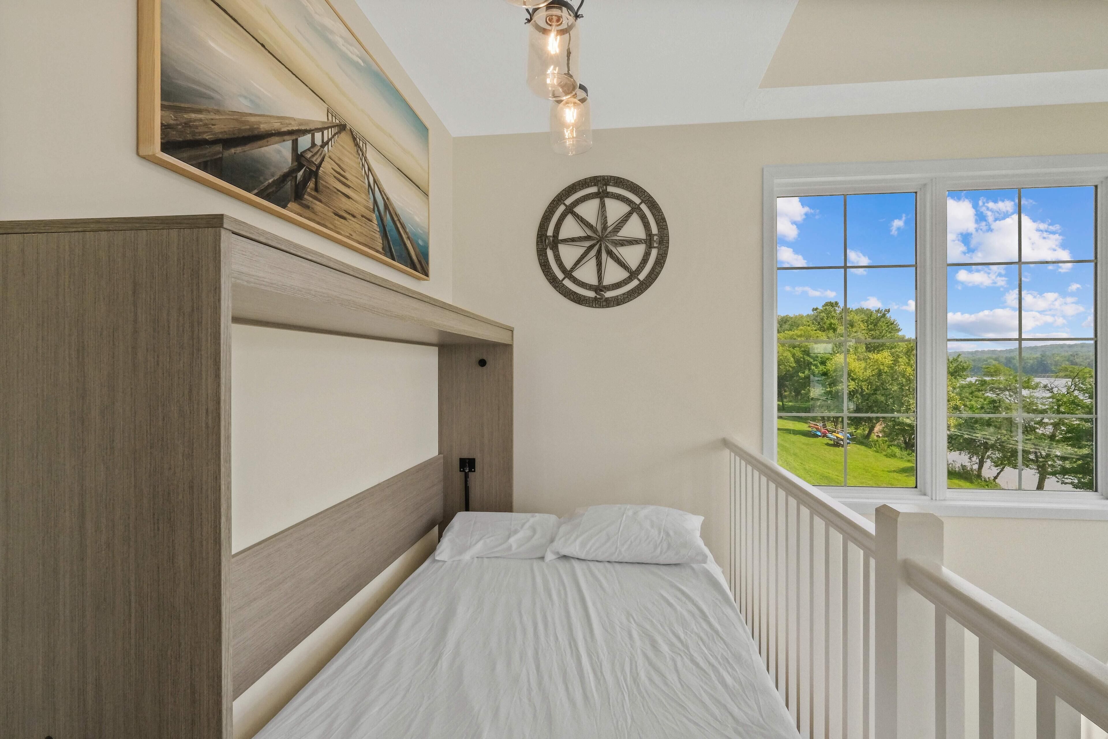 A bedroom with a wooden headboard, a compass wall decoration, a painting of a wooden dock, and a window with a view of greenery.