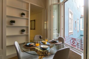 A dining area with a glass table, chairs, and a view of a blue building through a window. - Open Way - Mouzinho view Flat (Porto)