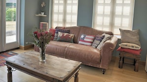 A living room with a brown leather sofa, a wooden coffee table, and a vase with flowers. There are two windows with blinds, a small table with a vase, and a chair with cushions.