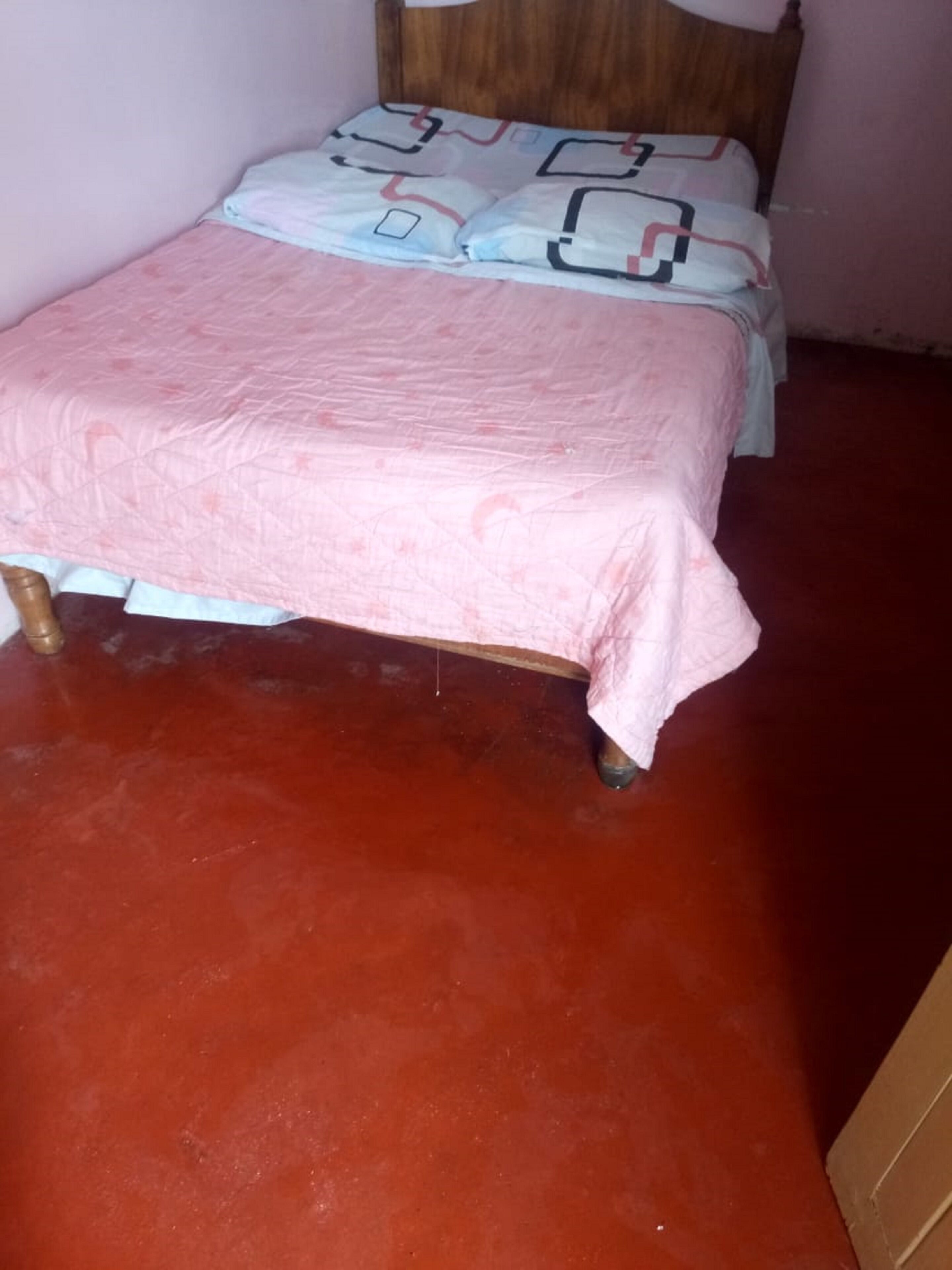 A single bed with a pink bedspread and a wooden headboard in a room with a red floor.