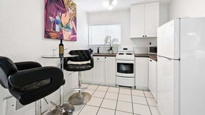 A compact kitchen with white cabinets, a white refrigerator, a stove, and a dining area with two black chairs and a small table. - MB Studio with Parking Sleeps 4 Steps to Sand (San Diego)