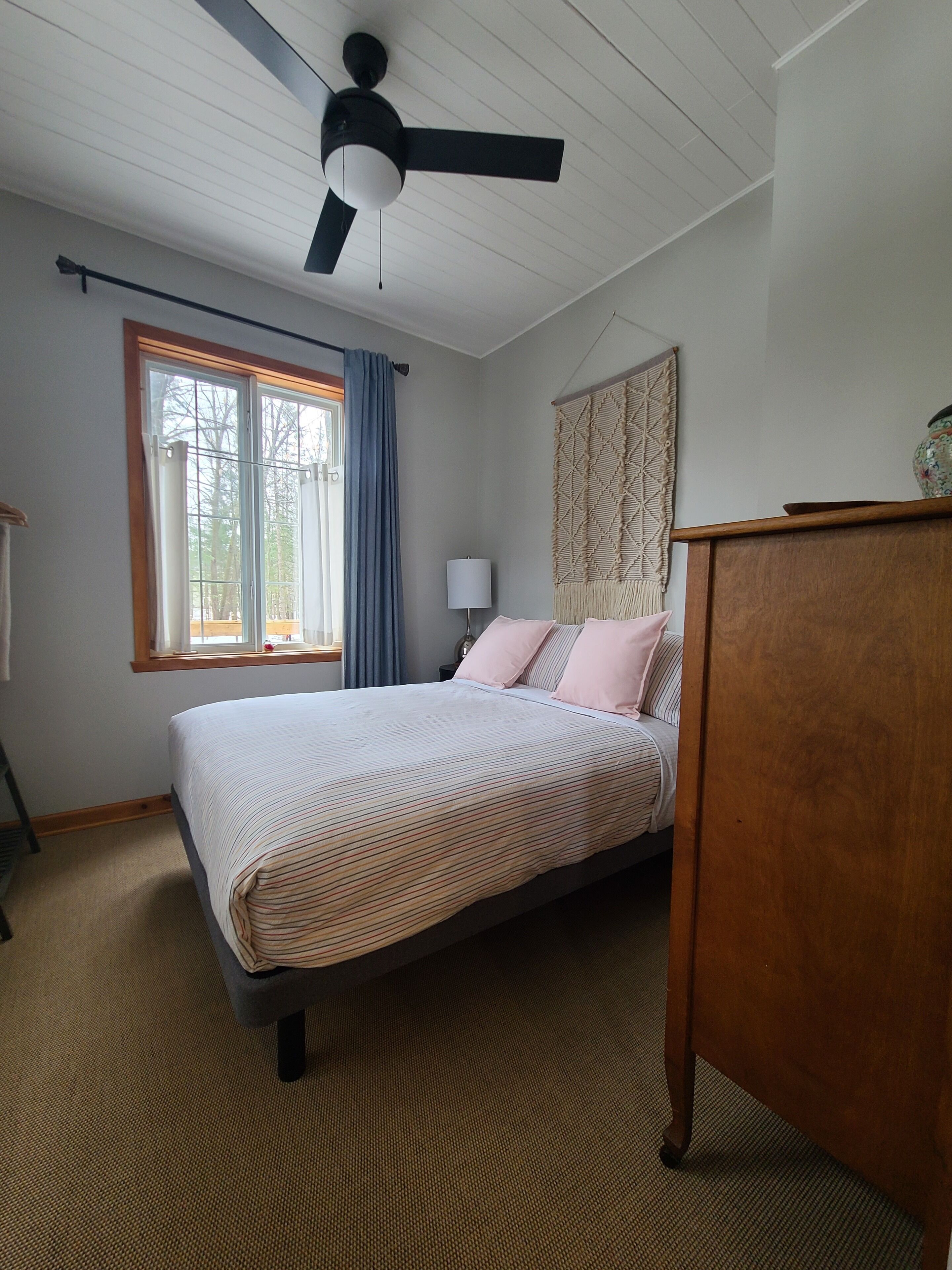 A bedroom with a bed, a ceiling fan, a window with curtains, a lamp, and a decorative wall hanging.