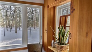 Un rincón de una habitación con una silla de madera, un ventanal con vistas a un bosque nevado y una estantería con una planta y dos botellas de vidrio.