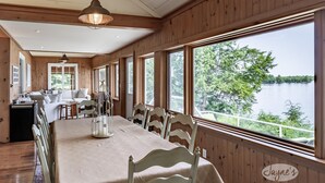 A dining area with a long table, white chairs, and a view of a lake. - Muskoka Paradise! Spacious & Inviting, Great Deck! Sleeps 16! Hot Tub! Bala! (Muskoka Lakes)