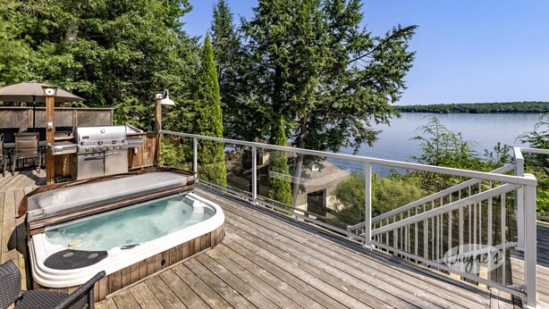 A wooden deck with a hot tub, barbecue, and outdoor furniture overlooking a lake. - Muskoka Paradise! Spacious & Inviting, Great Deck! Sleeps 16! Hot Tub! Bala! (Muskoka Lakes)