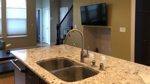 A kitchen with two stainless steel sinks, a granite countertop, a ceiling fan, and a mounted flat-screen TV. - Modern Living, relax and enjoy your stay! (Rancho cordova)