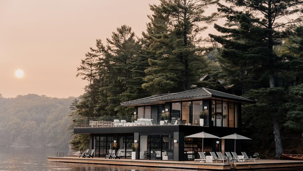 Una moderna casa flotante con terraza y sillones de descanso, rodeada de árboles y un lago.