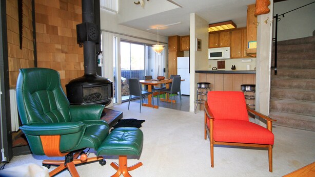 A spacious living area with a green leather chair, a red chair, and a green ottoman. A fireplace is visible in the background, and there is a kitchen area with wooden cabinets and a microwave. - Sierra Park Villas #24 (Mammoth Lakes)