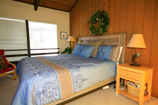 A bedroom with a bed, two bedside lamps, a wooden nightstand, a wicker basket, a red chair, and a window with blinds.