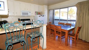 A kitchen with white cabinets, a microwave, and a refrigerator with magnets. There is a dining table with wooden chairs and a window with curtains. - Sierra Park Villas #16 (Mammoth Lakes)