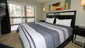 A bedroom with a large bed, a window with a view of trees, a painting on the wall, and a nightstand with a lamp. - Sierra Park Villas #03 (Mammoth Lakes)