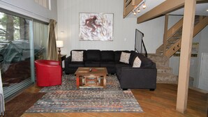A modern living room with a black sofa, a red ottoman, a wooden coffee table, and a colorful rug. There is a large abstract painting on the wall and a staircase leading to an upper level. - Sierra Park Villas #03 (Mammoth Lakes)