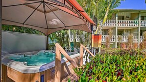 Zona de spa con piscina caliente de madera bajo un gran paraguas, rodeada de abundante vegetación y un edificio con balcones al fondo.