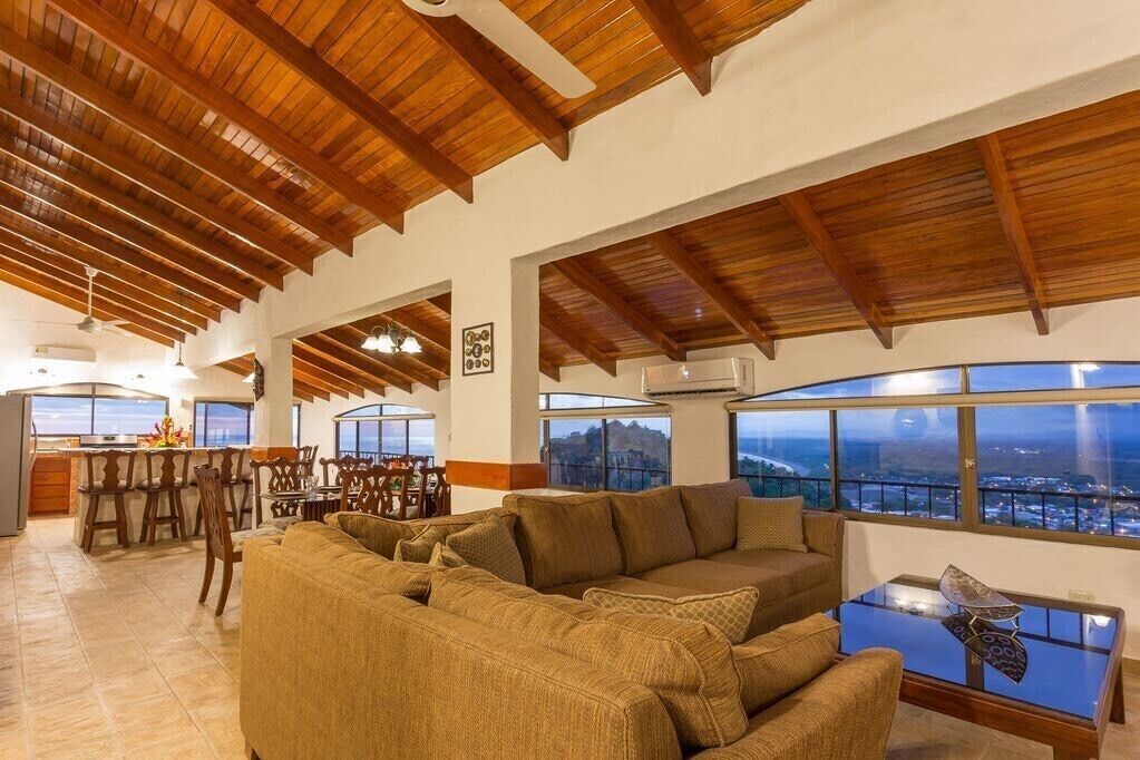 A spacious living area with a high ceiling, wooden beams, a large sofa, a dining table, and a view of the cityscape through large windows.
