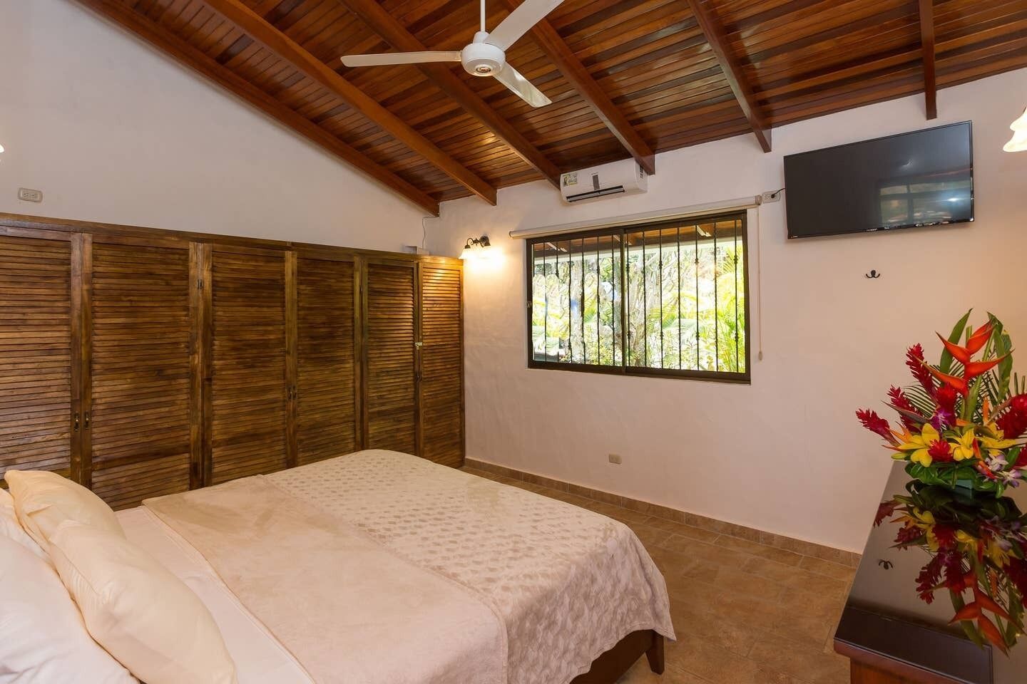 A bedroom with a bed, wooden wardrobe, a TV, and a window with a view of greenery.