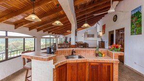 Une cuisine spacieuse dotée d’un grand îlot central, de poutres apparentes en bois au plafond et de grandes fenêtres offrant une vue sur l’extérieur.