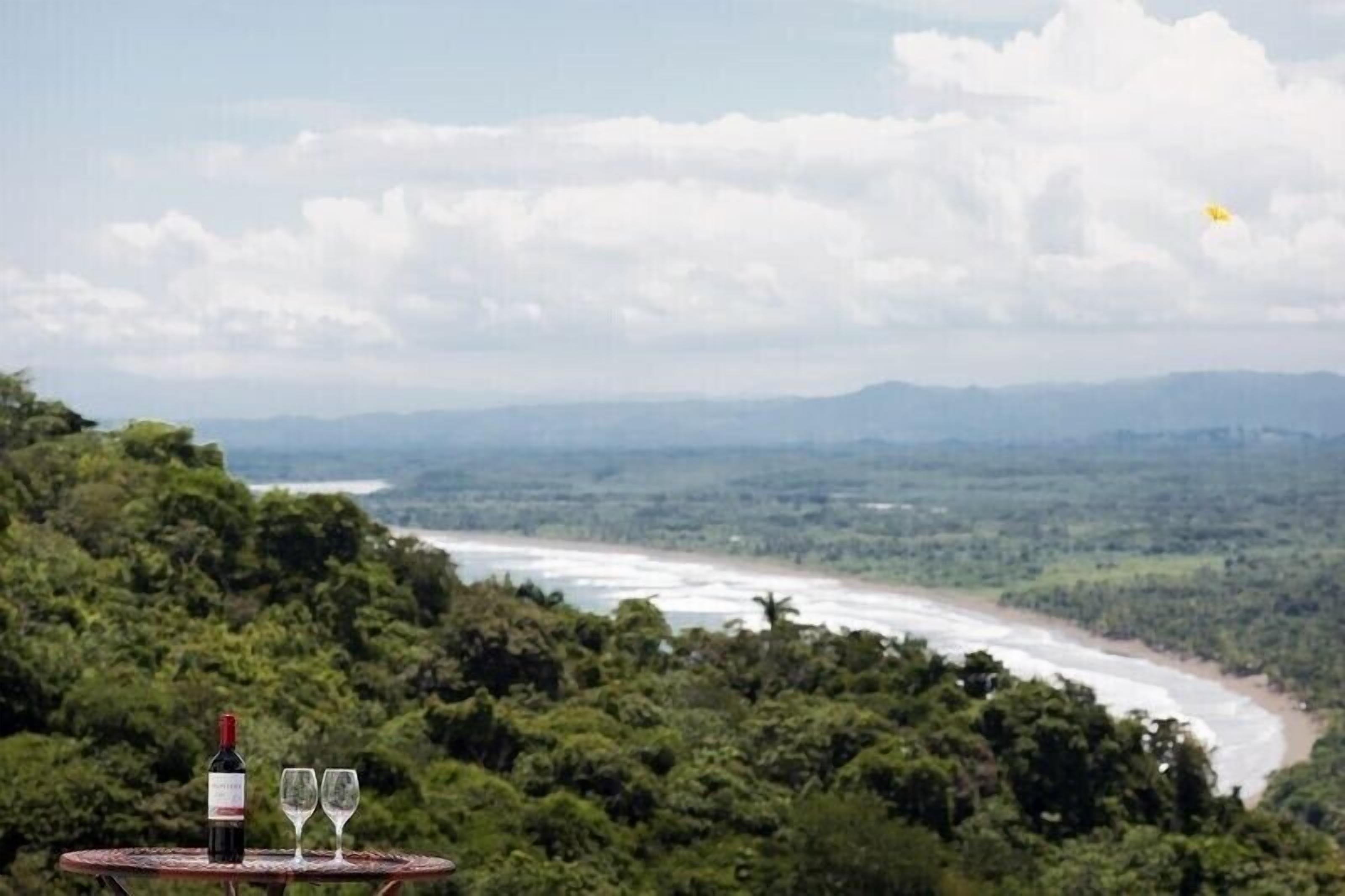 A table with a bottle and two glasses, overlooking a river and forested landscape.