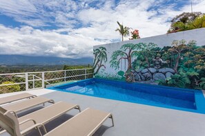 A swimming pool with a mural of tropical trees and rocks, surrounded by a white fence and beige lounge chairs. - Casa Moreno - Spectacular Views, Close to Beaches, Marina, Tours, Shopping! (Manuel Antonio)