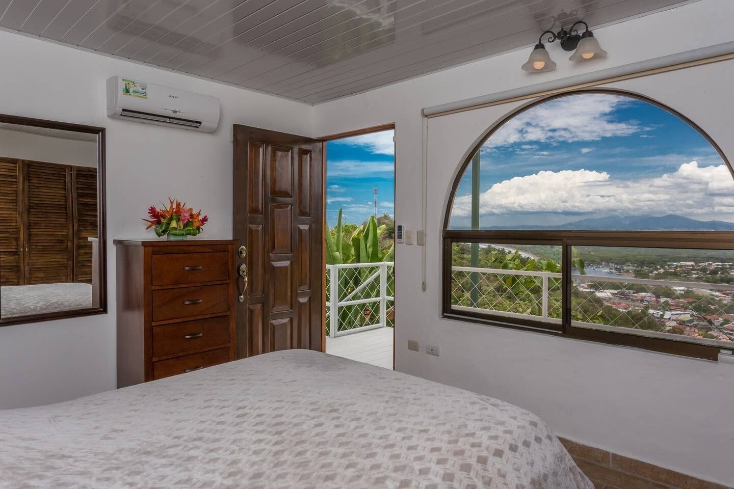 Un dormitorio con cama, cómoda, unidad de aire acondicionado y ventana con vista a un paisaje urbano y montañas.