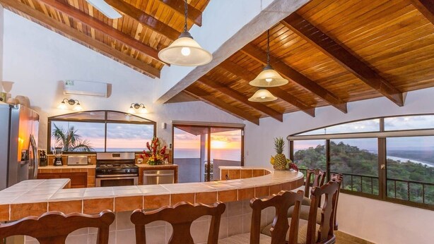Une cuisine spacieuse dotée d’un grand îlot central, un coin repas avec des chaises en bois et une vue sur le paysage grâce à de grandes fenêtres.