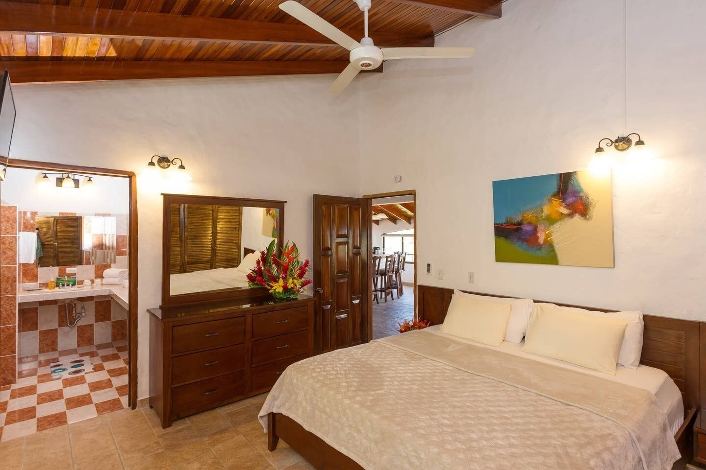 Une chambre d’hôtel avec un lit, une commode, une coiffeuse avec un miroir, un ventilateur de plafond et un tableau au mur.