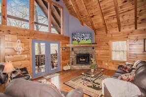 A log cabin interior with a stone fireplace, wooden ceiling beams, a large TV, and a cozy seating area.