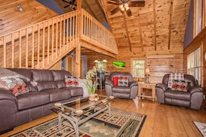 A spacious living room with a wooden ceiling, a balcony, leather sofas, a glass coffee table, and a fireplace.