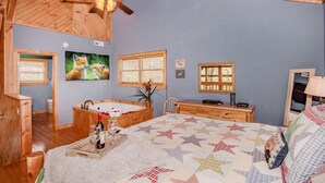Una camera da letto con soffitto in legno, una ventola a soffitto, un letto grande con copripiumino a motivi stellari, un comò in legno, un comodino, una TV e una finestra con persiane.