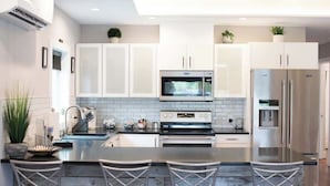 A modern kitchen with white cabinets, a central island with bar stools, stainless steel appliances, and a tiled backsplash. - Downtown Solar Home w/ Hot Tub & Billiards (Geneva)