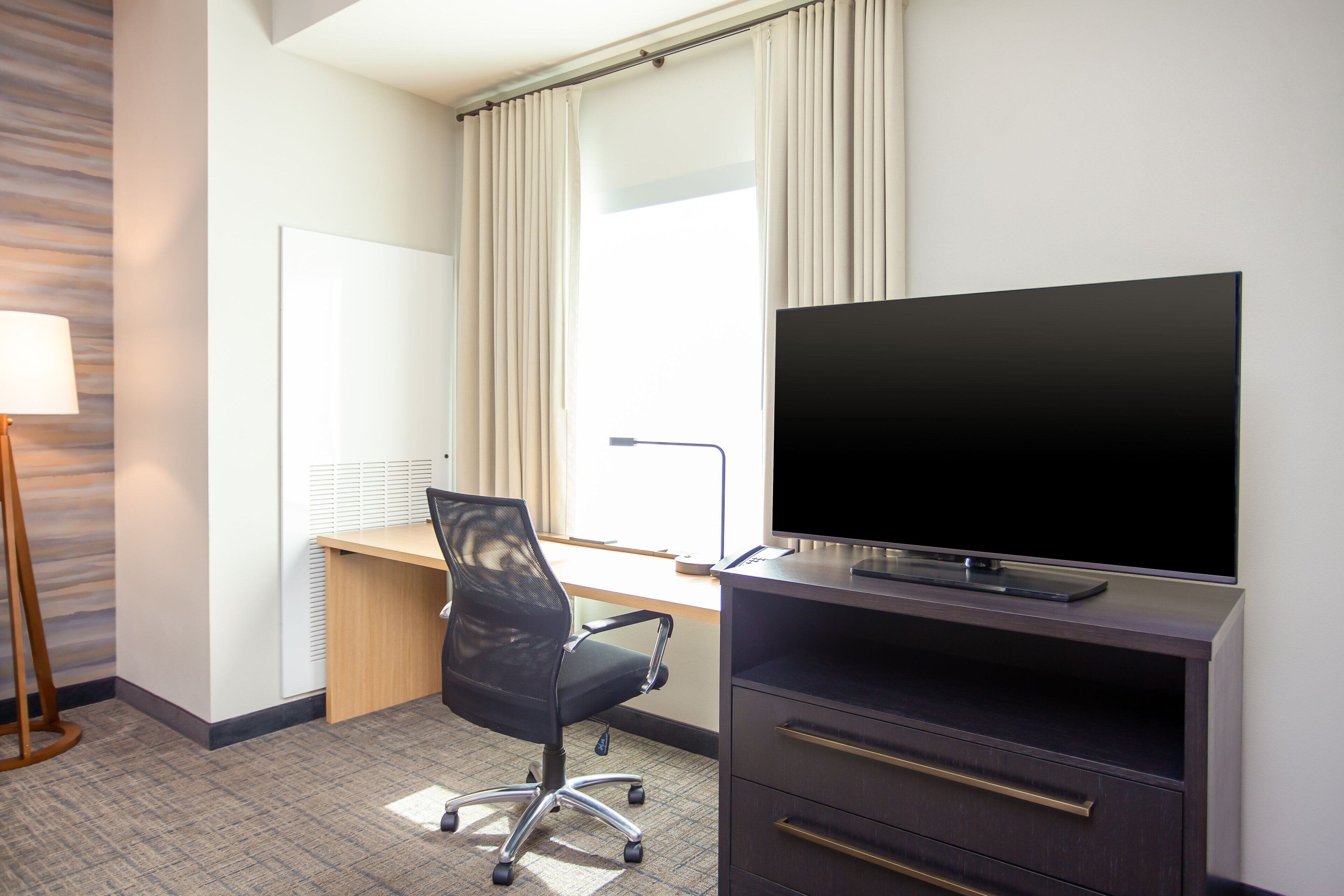 A hotel room with a desk, chair, TV, and a lamp.