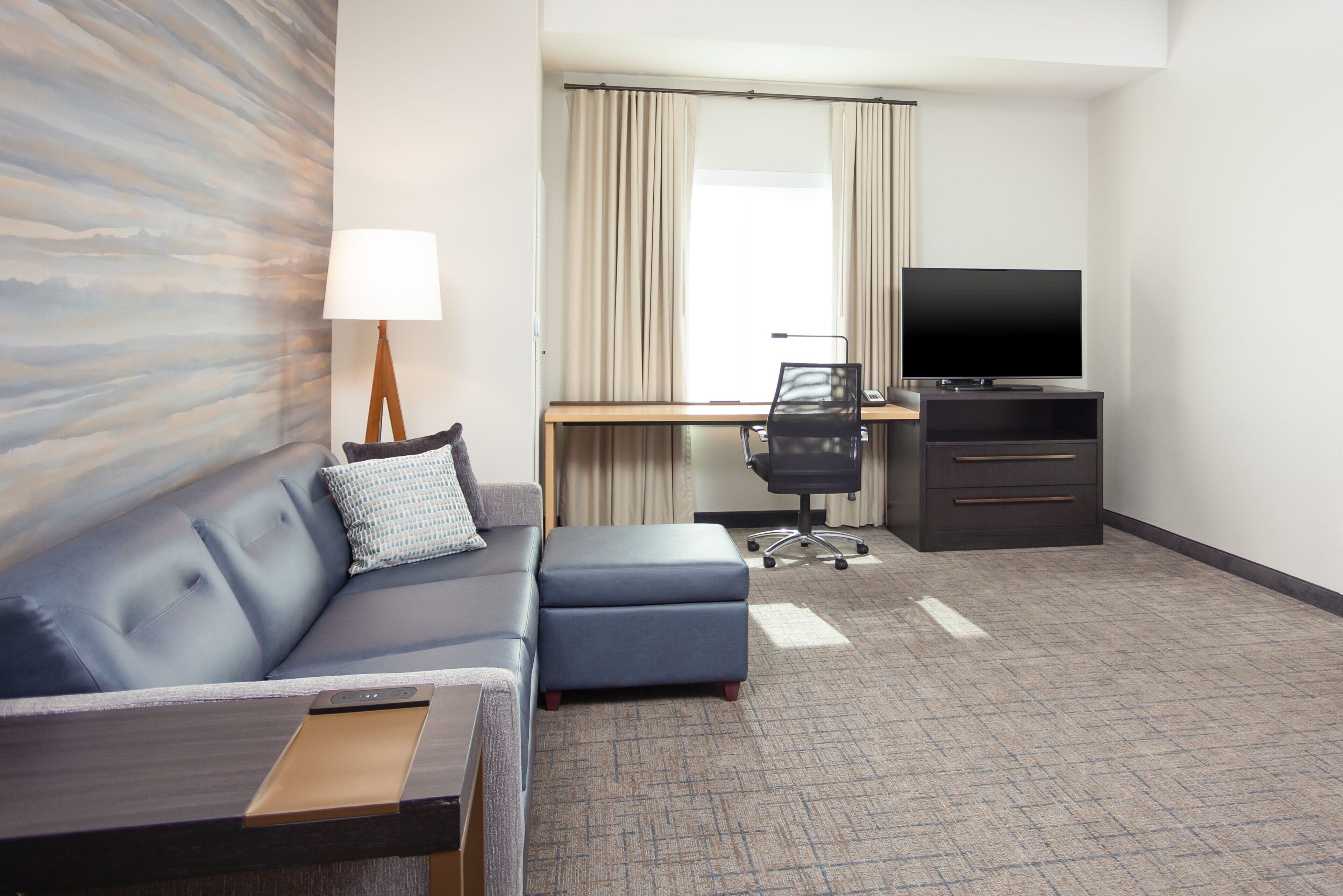 A hotel room with a sofa, a desk with a computer, a television, and a lamp.