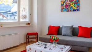 Un soggiorno con un divano grigio, cuscini rossi e grigi, un tavolo con tovaglia floreale e un vaso di fiori, una finestra con vista sugli edifici e un quadro astratto colorato appeso al muro.