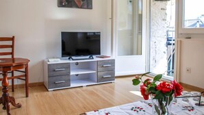 Una stanza con una TV su un mobile, una sedia di legno, un tavolino con un vaso di fiori e una porta scorrevole in vetro che dà su uno spazio esterno.