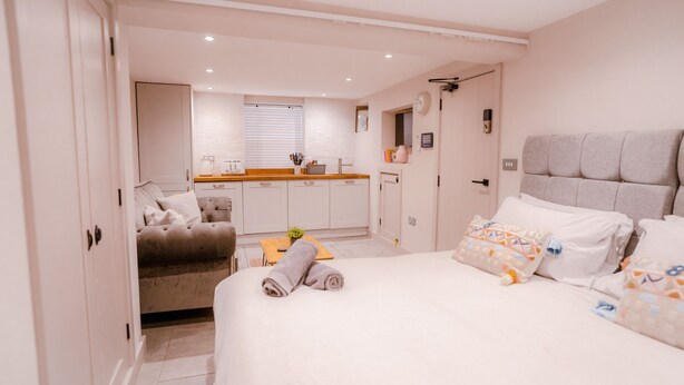 Ein Schlafzimmer mit einem Bett, einem Sofa und einem kleinen Tisch. Im Hintergrund ist eine Küchenzeile mit einem Fenster und einer Spüle zu sehen.