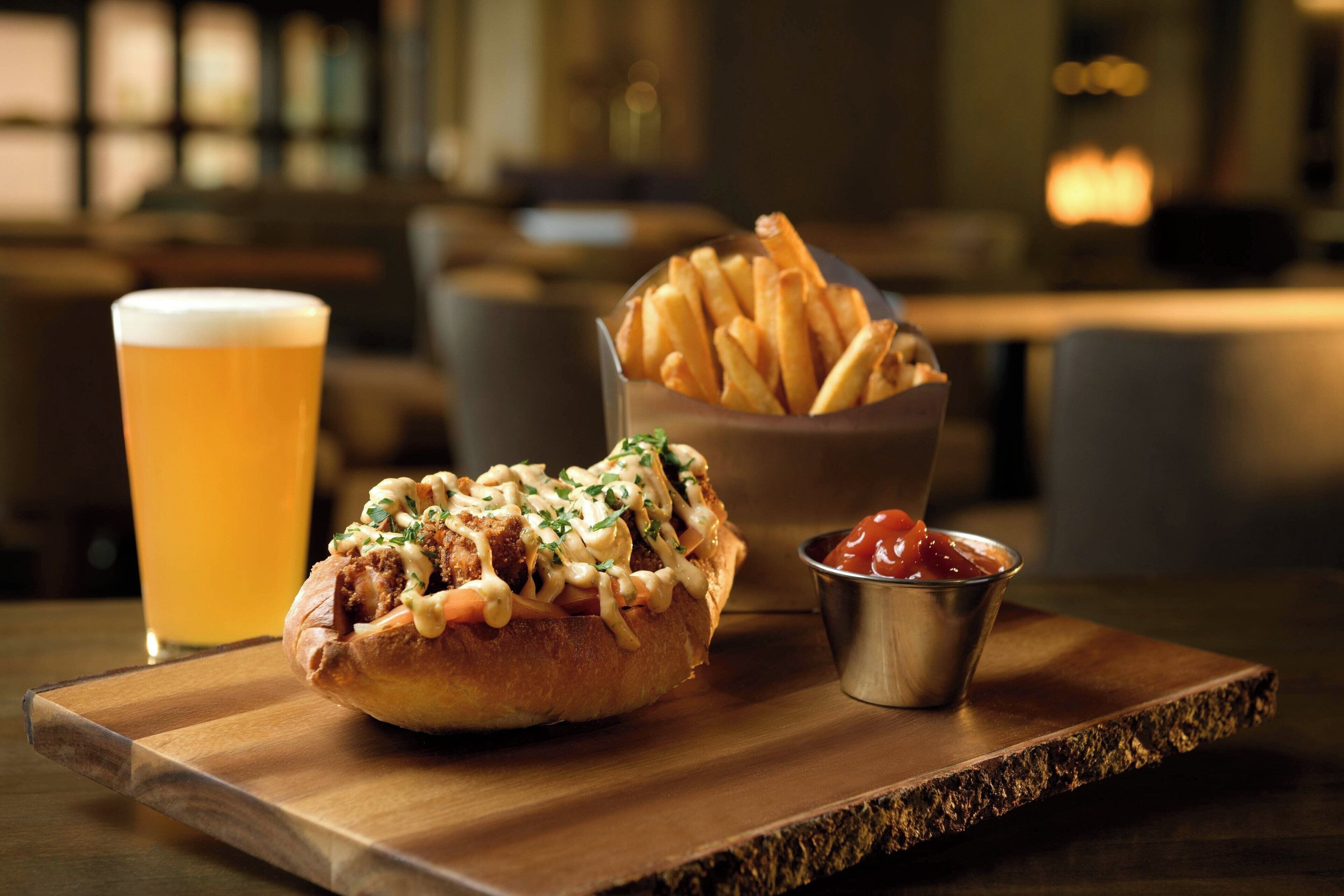 A meal consisting of a burger, fries, a glass of beer, and a small bowl of ketchup on a wooden board.