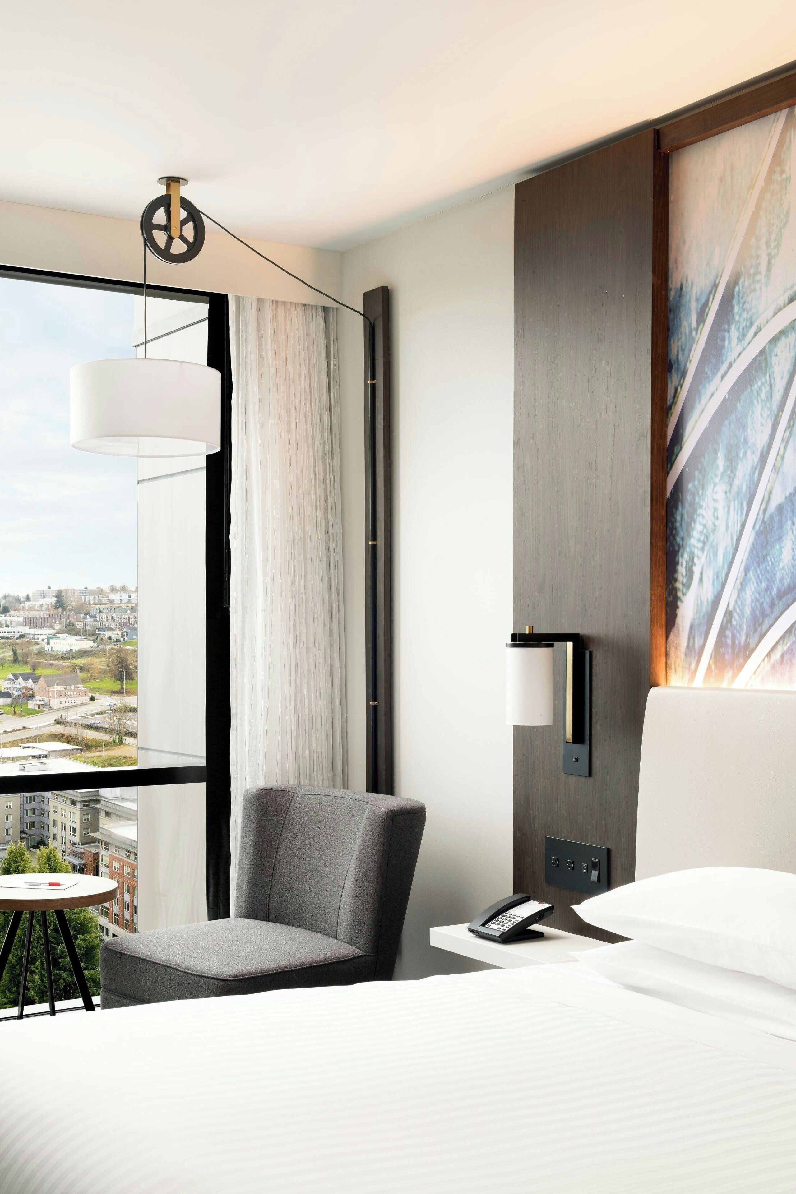 Ein Hotelzimmer mit einem Bett, einem Sessel, einem Schreibtisch mit Telefon, einer Wandlampe und einem Gemälde.