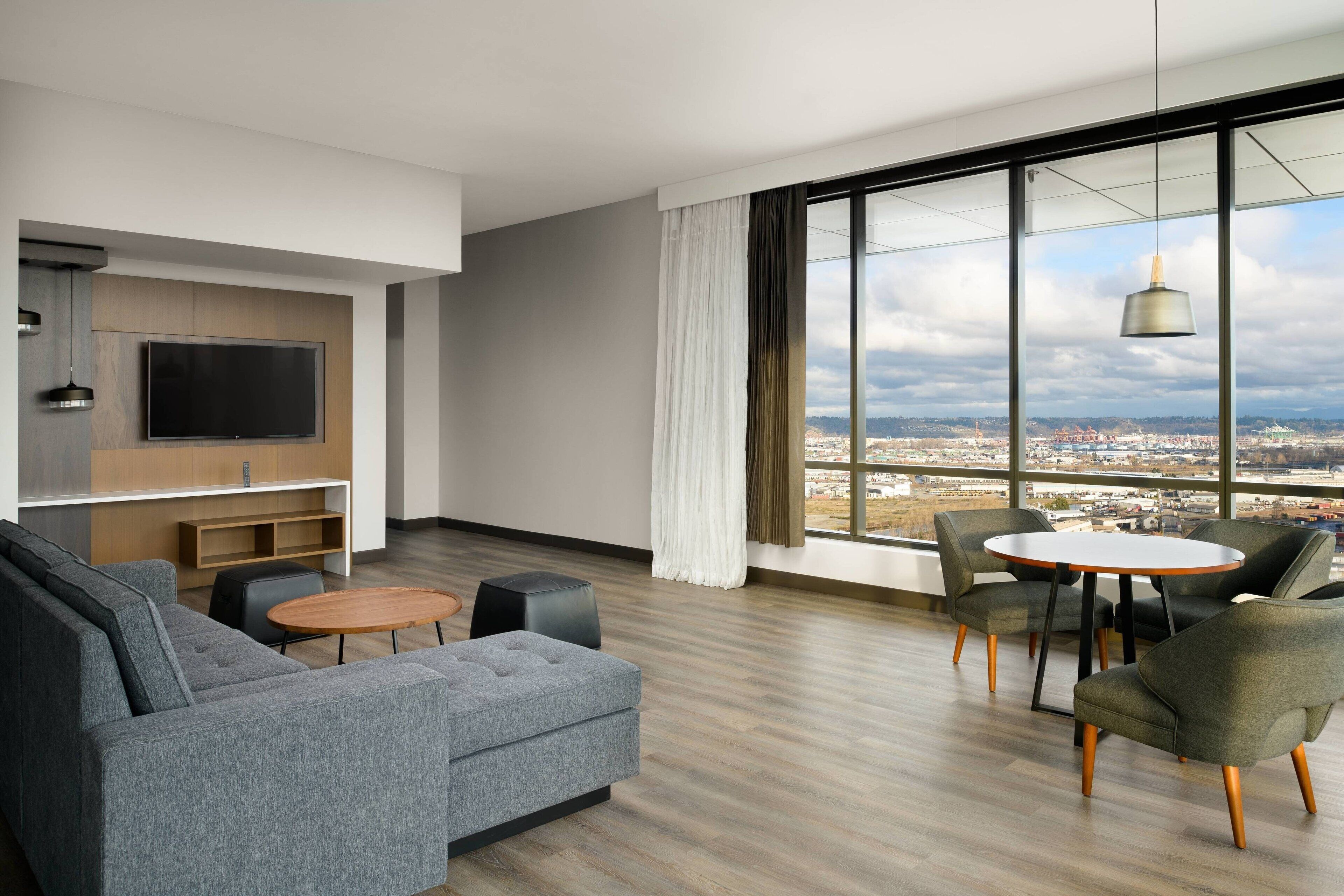 A modern living room with a sofa, a coffee table, and a dining table with chairs. There is a flat-screen TV mounted on the wall and a large window offering a city view.