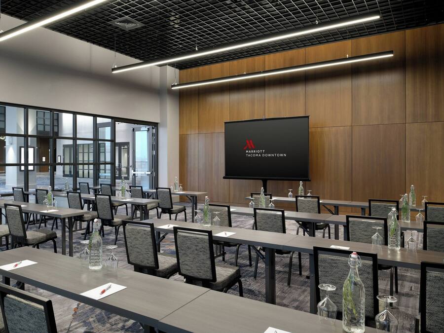 A conference room with multiple tables and chairs, a large screen displaying the Marriott logo, and a view of the outside through large windows.