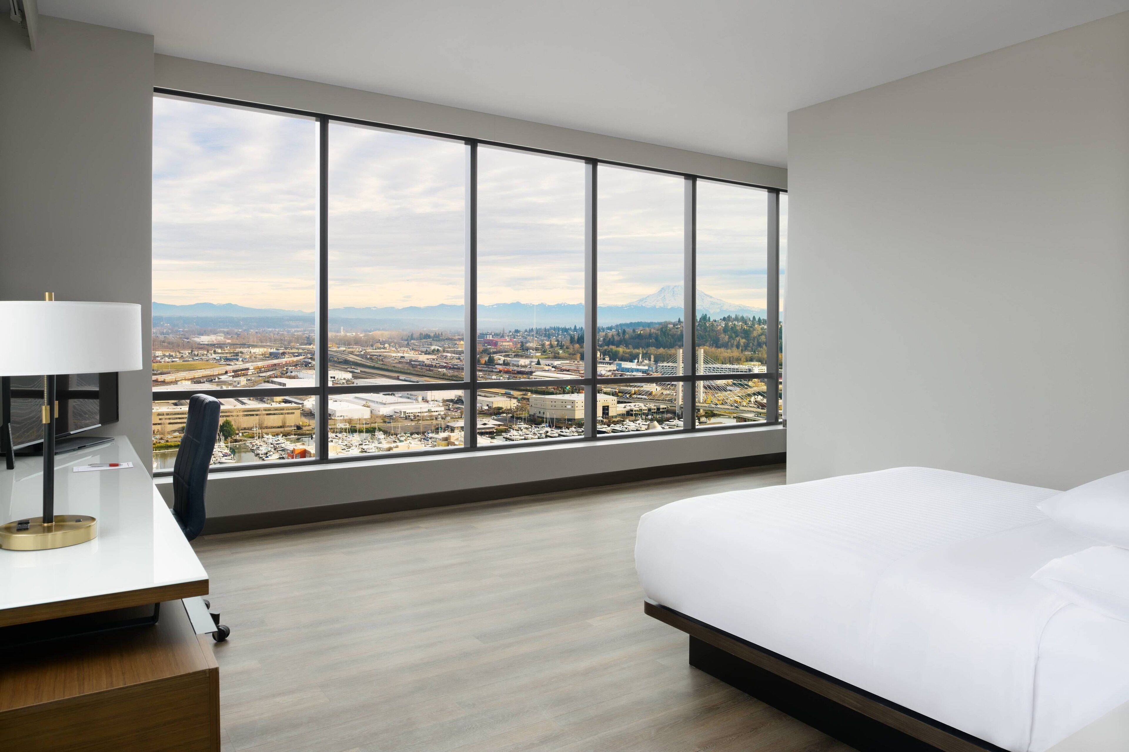 Quarto de hotel com uma janela ampla que oferece vista para a cidade, uma cama, uma escrivaninha com luminária e uma cadeira.