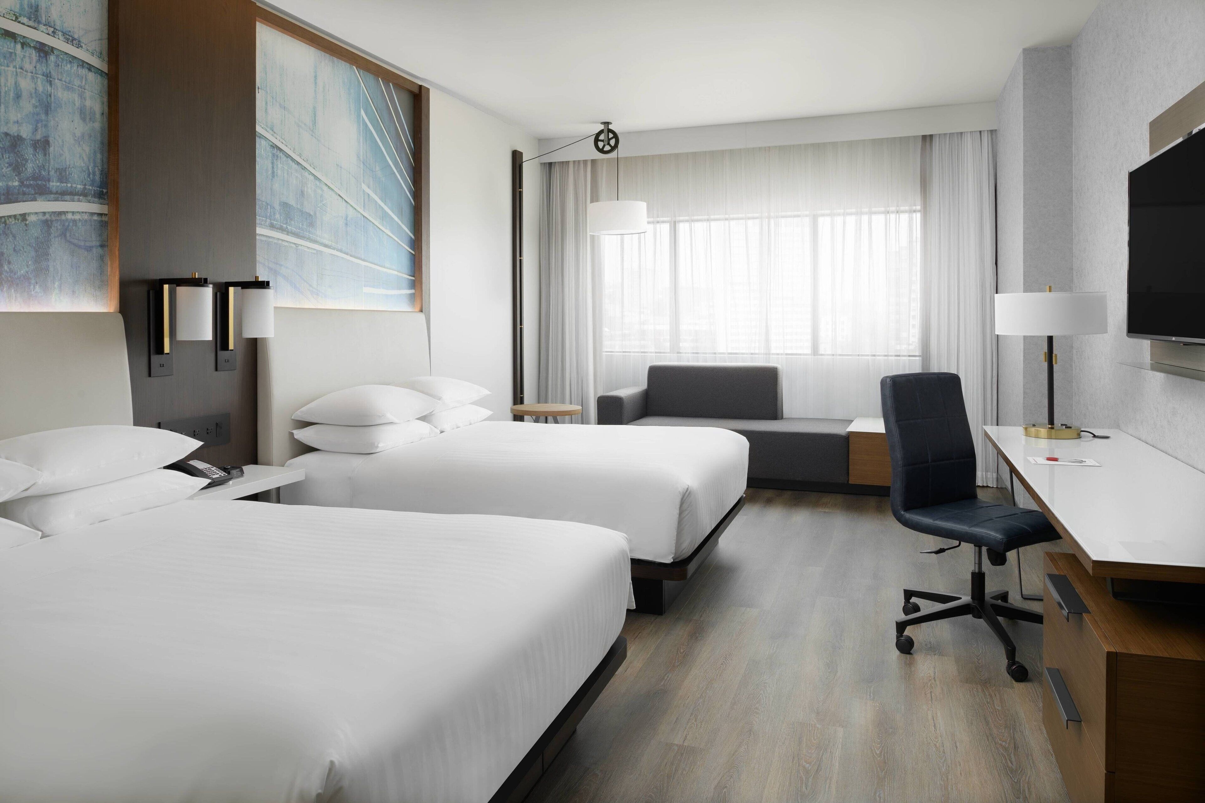 Ett hotellrum med två sängar, ett skrivbord, en stol, en tv och ett stort fönster med gardiner.