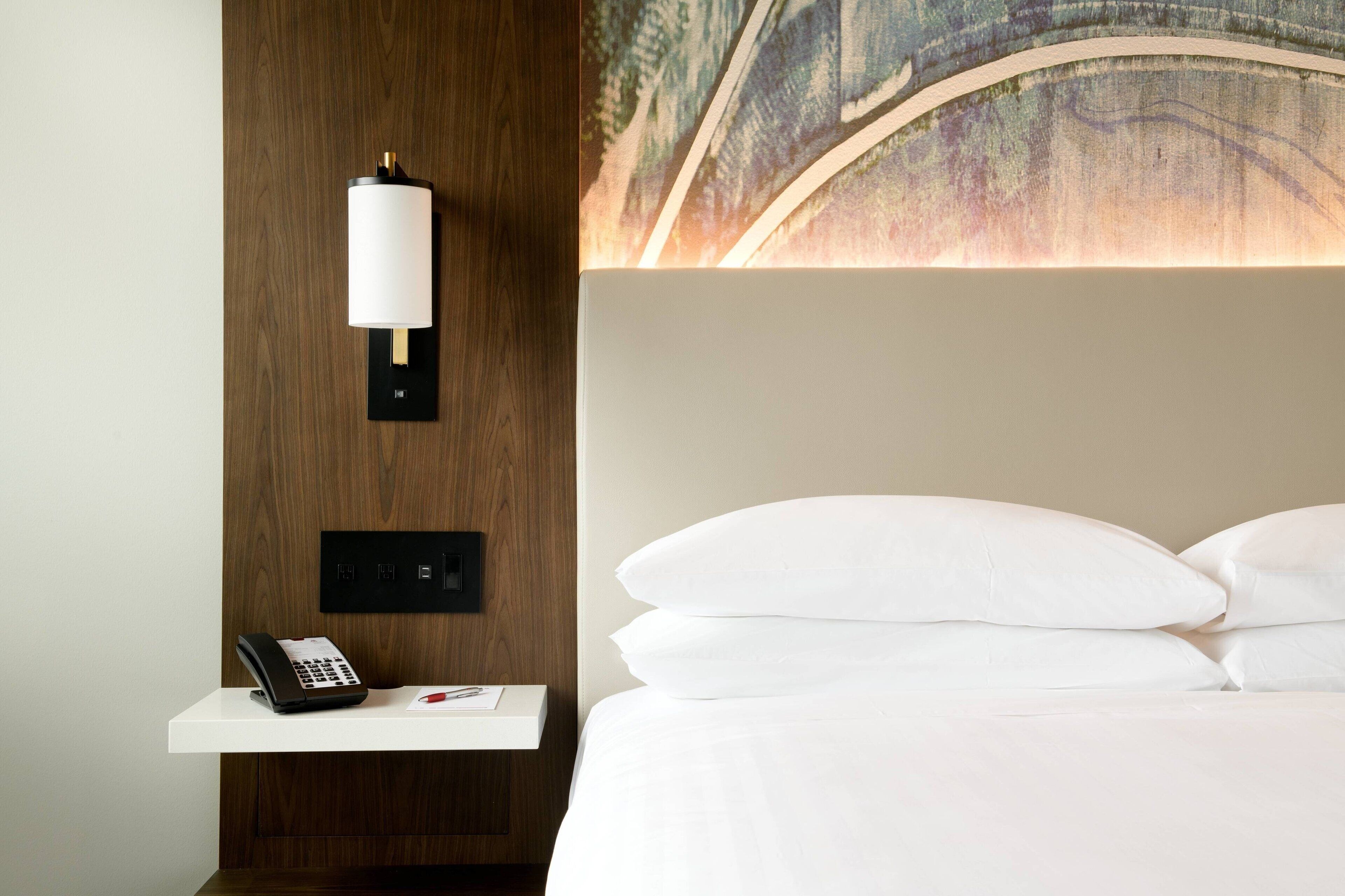 Une chambre d’hôtel avec un lit, des oreillers blancs, une table de chevet avec un téléphone et une lampe fixée au mur.