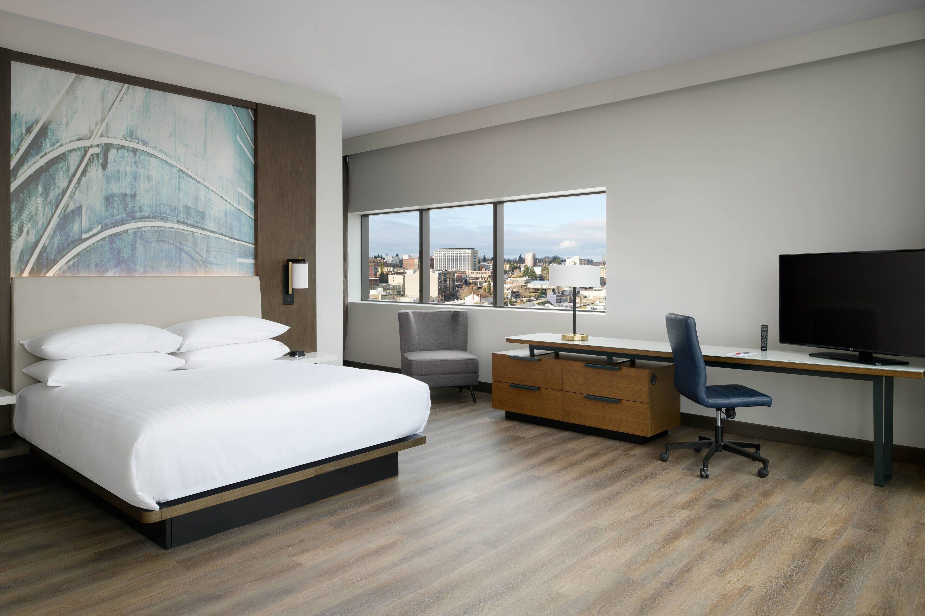 Une chambre d’hôtel moderne dotée d’un grand lit, d’un bureau avec une chaise, d’une télévision et offrant une vue sur la ville grâce à de grandes fenêtres.
