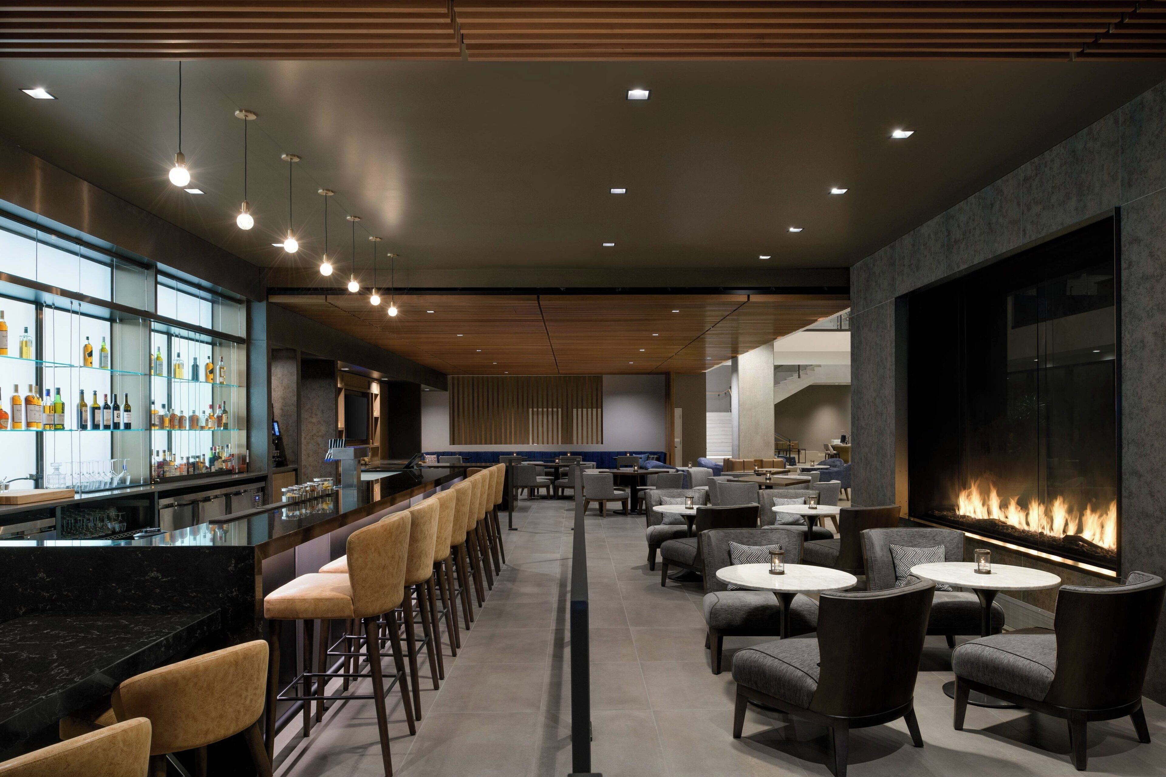 A modern bar area with a long counter, bar stools, and a variety of bottles. There is a seating area with round tables and chairs, and a large wall-mounted screen.