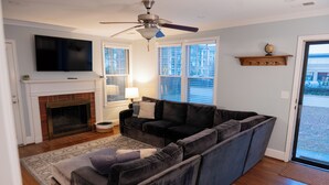 Ein Wohnzimmer mit einem Ecksofa, einem Flachbildfernseher über einem Kamin, einem Deckenventilator und großen Fenstern.
