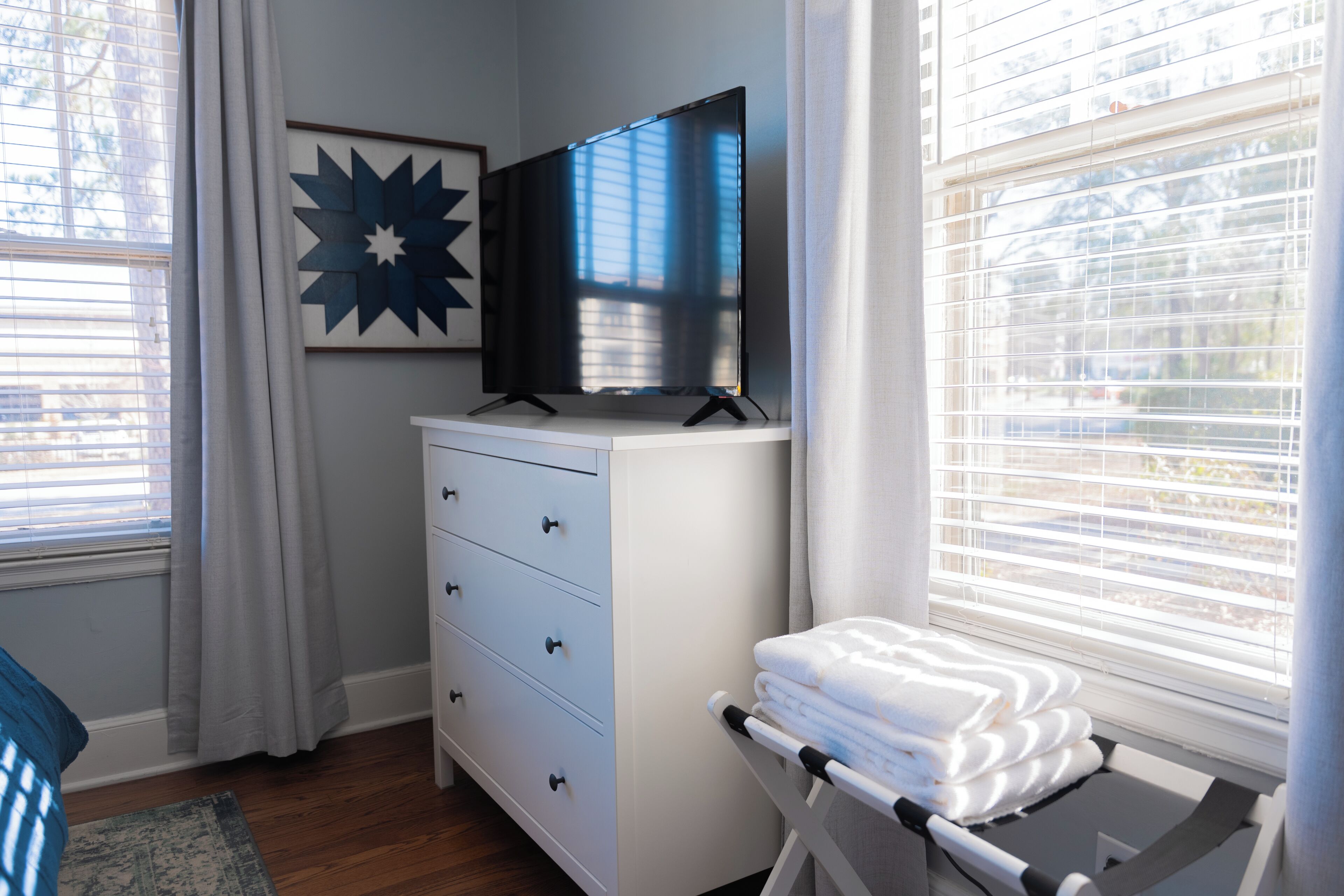 A bedroom with a TV on a dresser, a window with blinds, and a bed with a blue pillow.