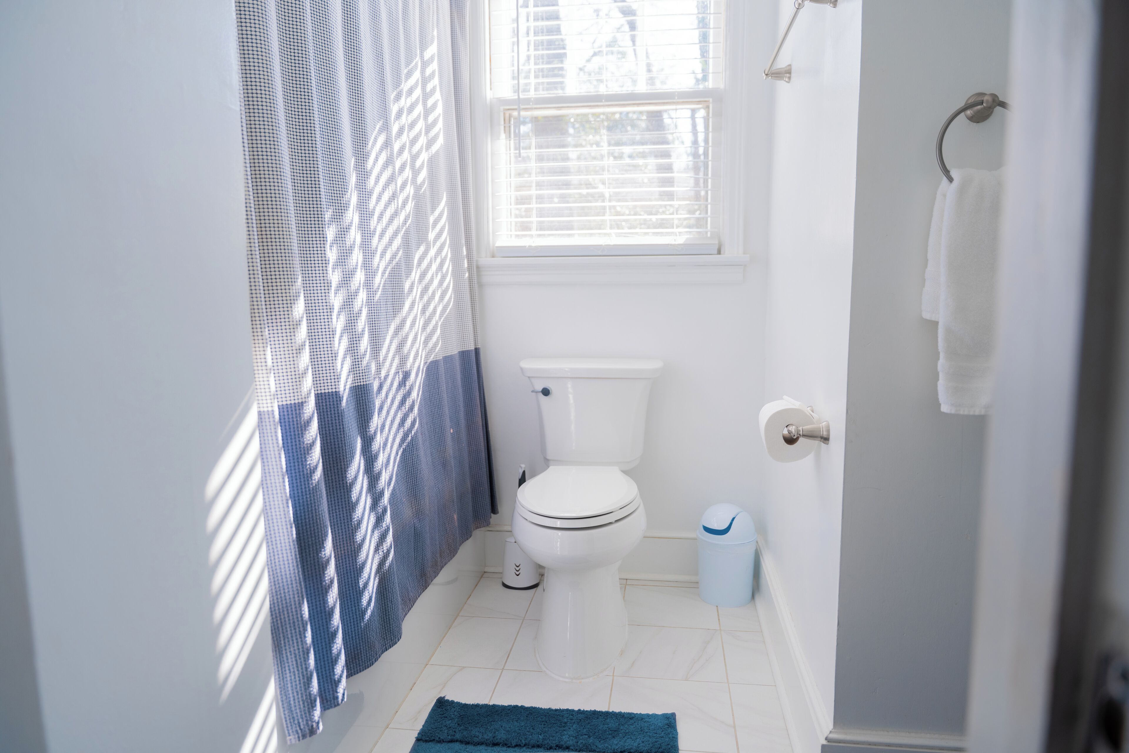 A bathroom with a toilet, a trash bin, a towel rack, and a window with blinds.